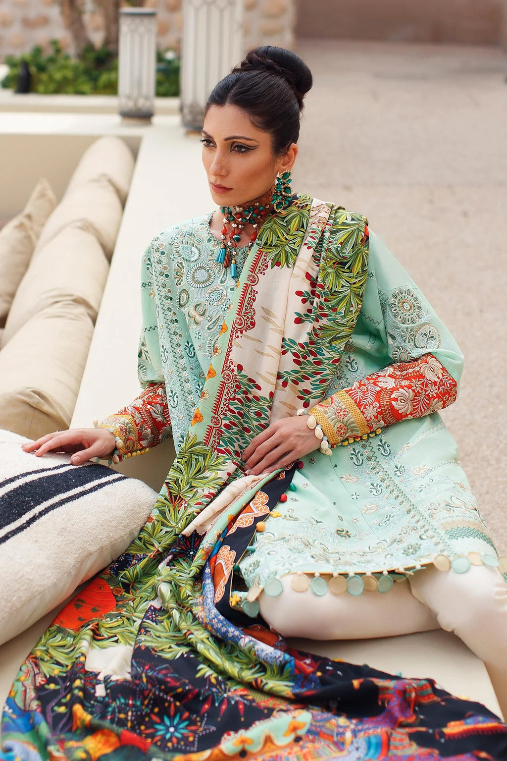 A woman seated outdoors wearing a traditional embroidered outfit with a mint green kurta, multicolored printed dupatta, and cream pants. The setting includes cushions and a stone architectural background.