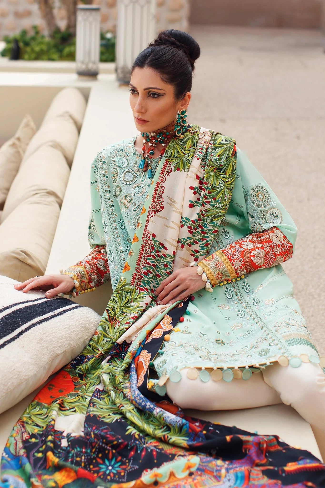 A woman seated outdoors wearing a traditional embroidered outfit with a mint green kurta, multicolored printed dupatta, and cream pants. The setting includes cushions and a stone architectural background.