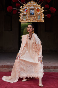 Woman in a beige embroidered outfit standing on a red carpet with decorative background
