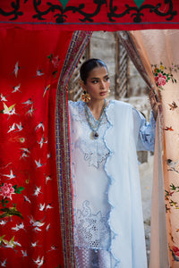 Woman in a light blue traditional outfit standing behind a decorative red and pink screen with floral patterns.