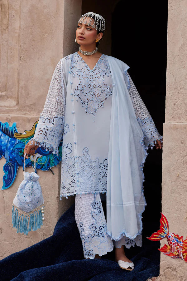 Woman in traditional white and blue embroidered outfit with a headscarf, standing in front of a decorative wall.
