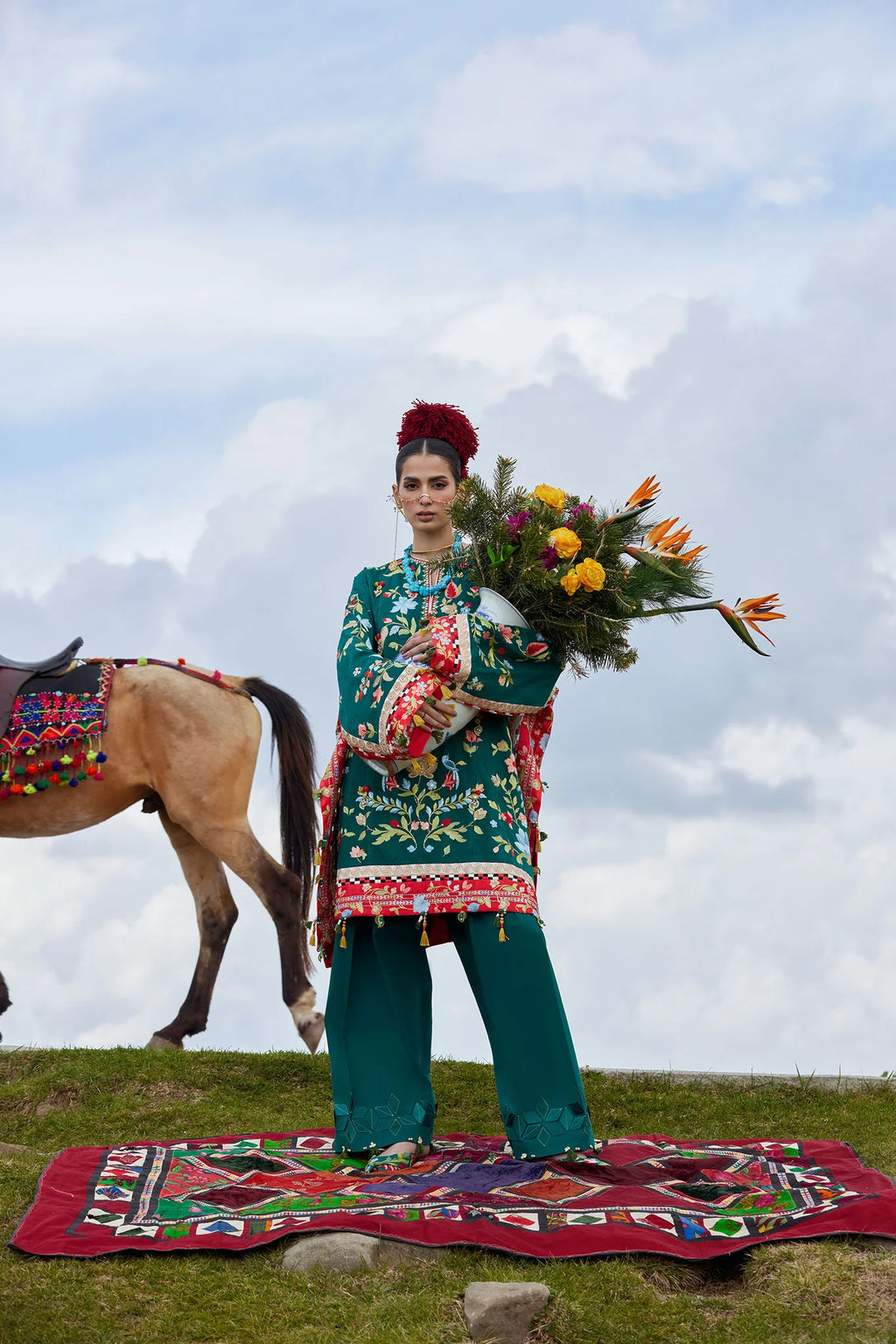 Woman in traditional attire holding flowers with a horse and colorful rug in the background