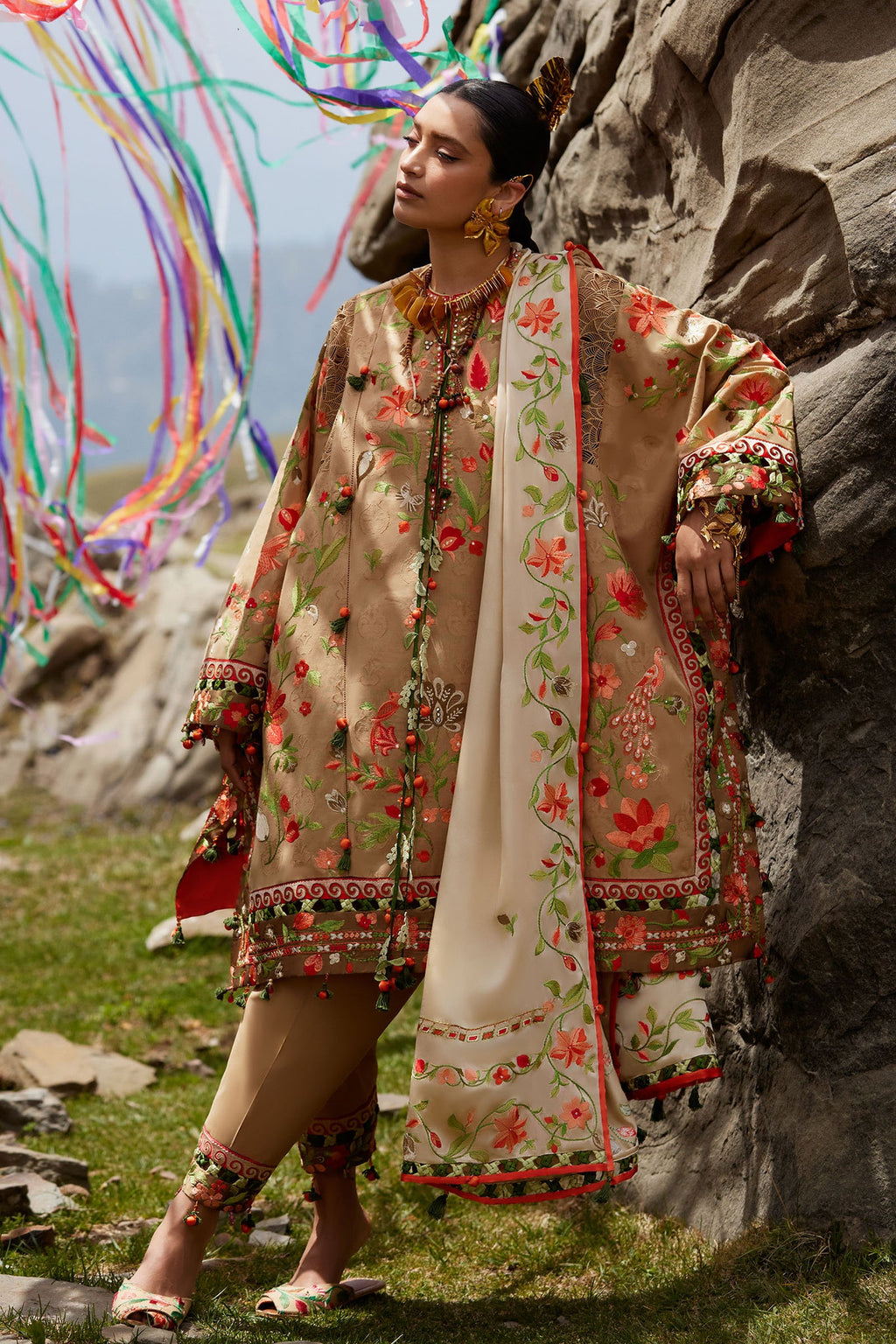 Woman in traditional embroidered outfit standing against a rocky background with colorful ribbons.