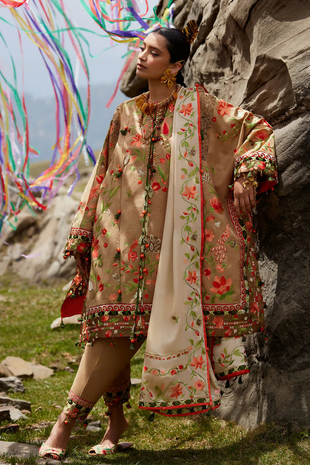 Woman in traditional embroidered outfit standing against a rocky background with colorful ribbons.