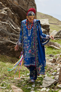 Person in traditional blue embroidered outfit with colorful accessories standing in a rocky, mountainous landscape.