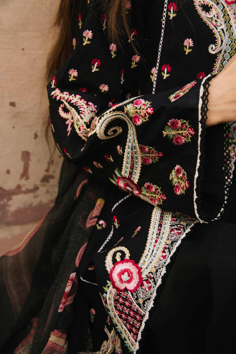 Close-up of a black embroidered traditional garment with floral and paisley motifs in pink, red, green, and beige, worn by a person whose arm and hand are visible.
