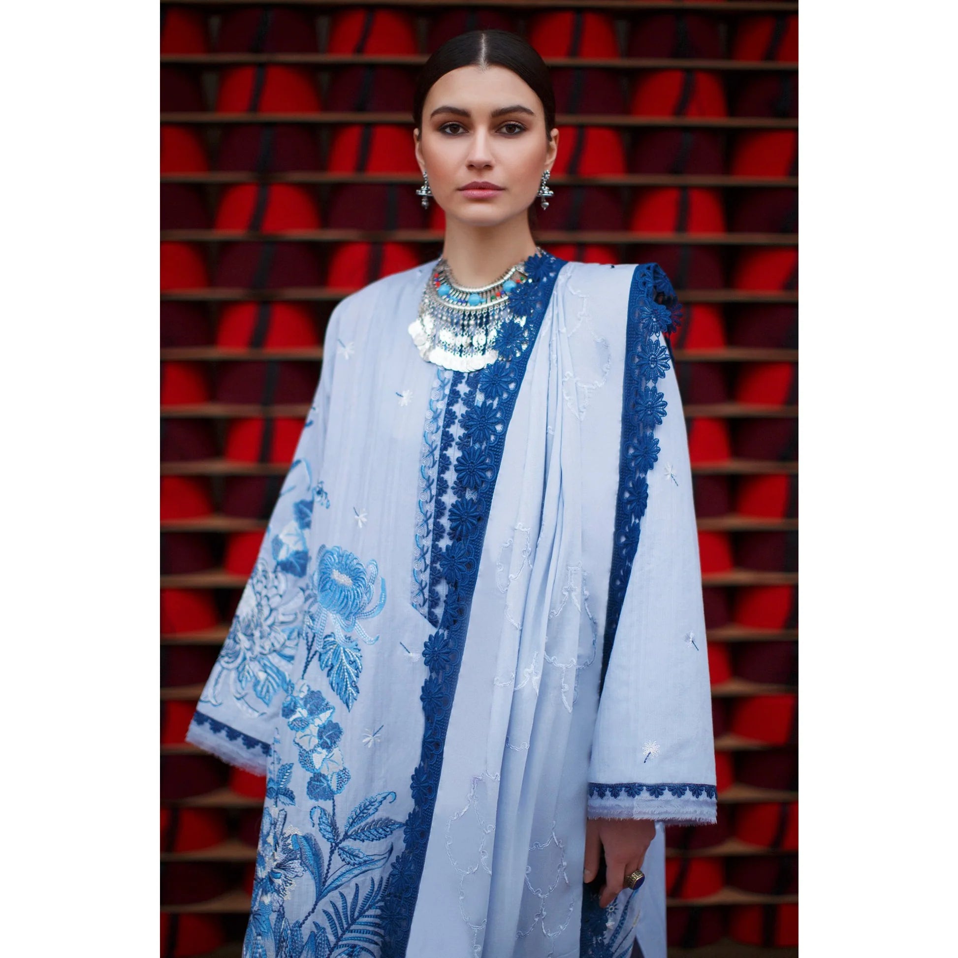 A woman modeling a traditional light blue and navy embroidered outfit with a matching dupatta, set against a background of red stadium seating.