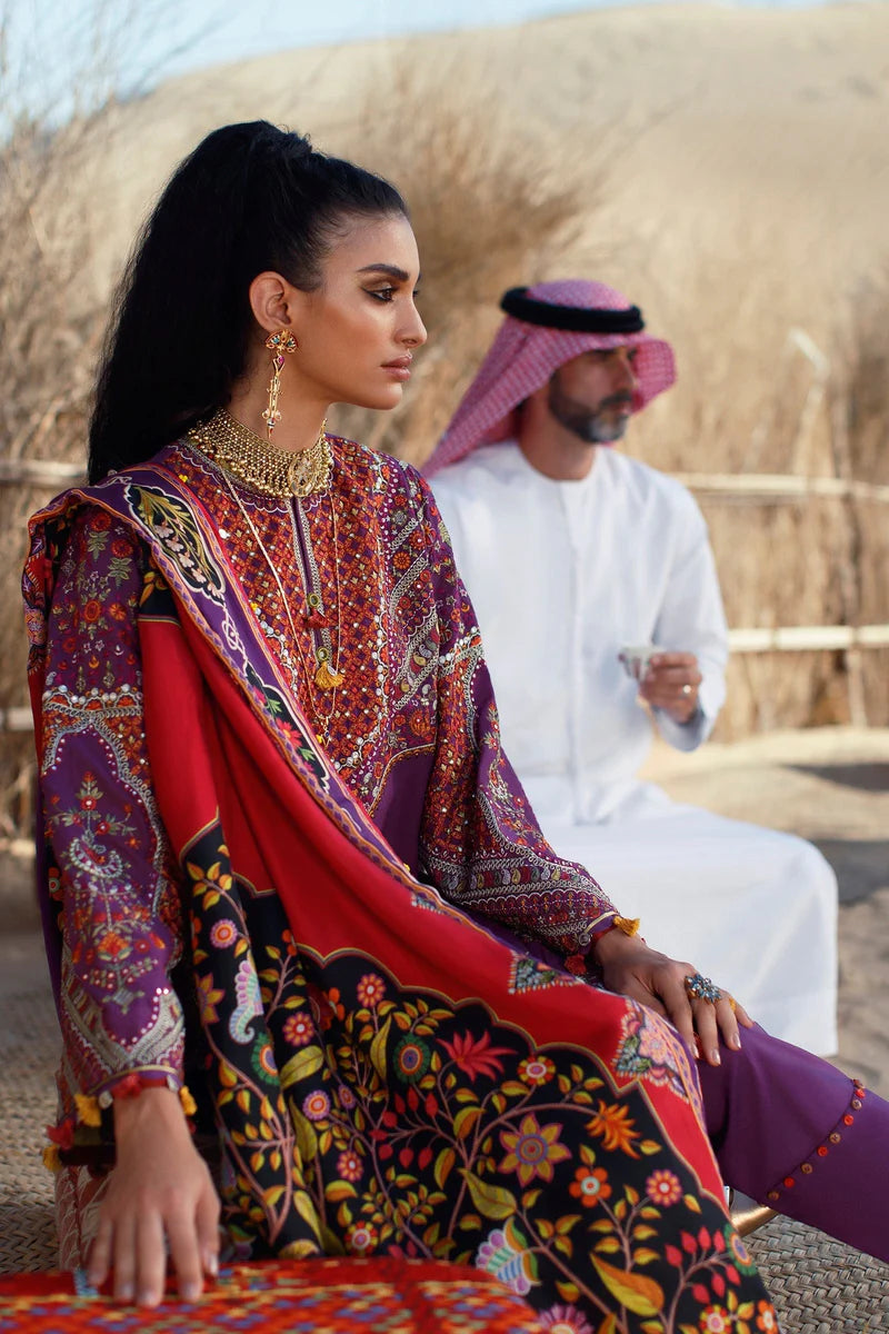 A woman in a traditional South Asian outfit with a red, purple, and black floral printed salwar kameez and dupatta, seated outdoors in a desert setting with a man in a white thobe and pink headscarf in the background.