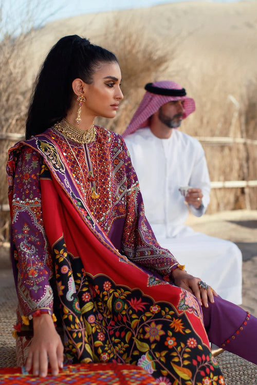 A woman in a traditional South Asian outfit with a red, purple, and black floral printed salwar kameez and dupatta, seated outdoors in a desert setting with a man in a white thobe and pink headscarf in the background.