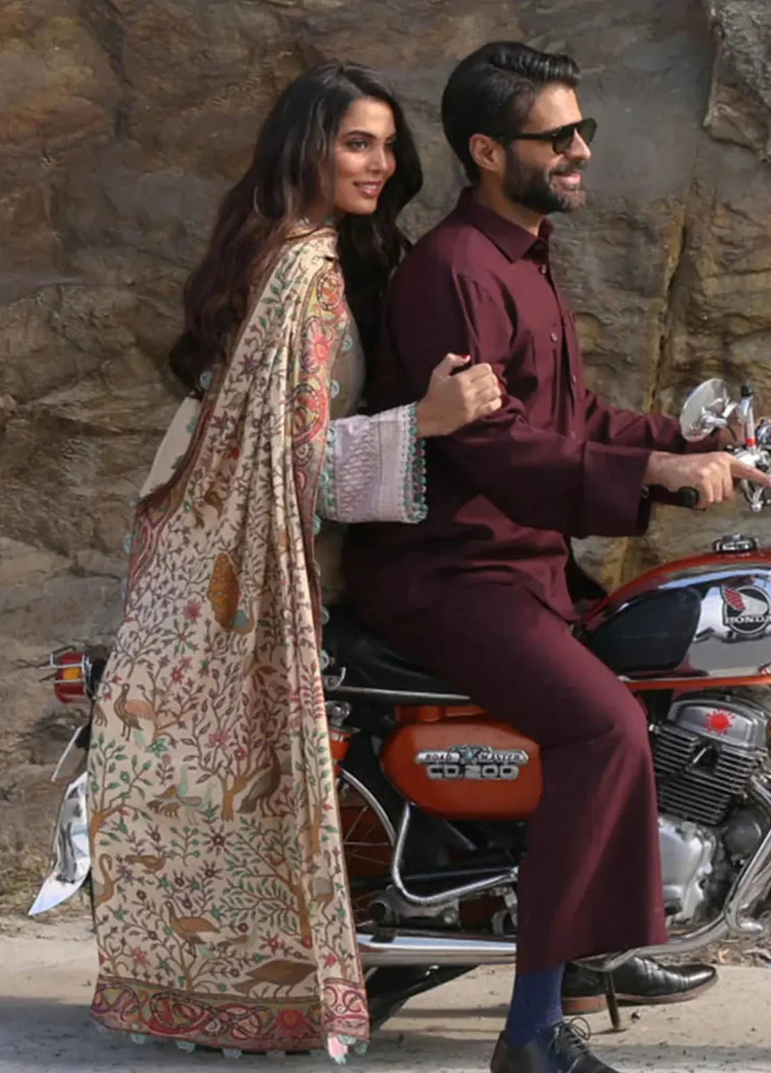 A woman in a beige and red patterned shawl and a man in a maroon traditional outfit riding a motorcycle against a rocky background.