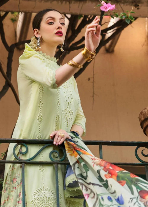 A woman wearing a light green embroidered traditional outfit with a floral printed dupatta, posing on a balcony with a pink flower in her hand.