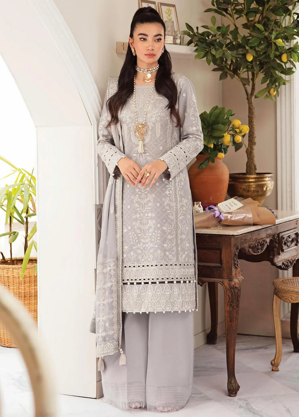 A woman models a light gray three-piece traditional outfit with intricate white embroidery, matching wide-leg trousers, and a draped dupatta, set against an elegant indoor background with a wooden table and potted plants.