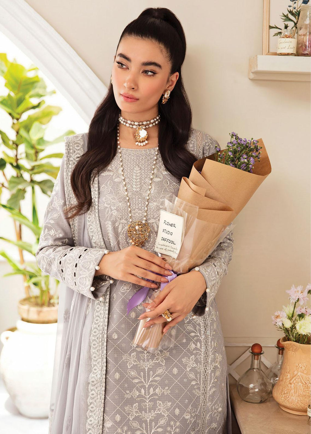 A woman models a light gray embroidered traditional outfit with matching long sleeves and a draped outer layer, holding a brown paper-wrapped bouquet in a bright indoor setting with plants and home decor.