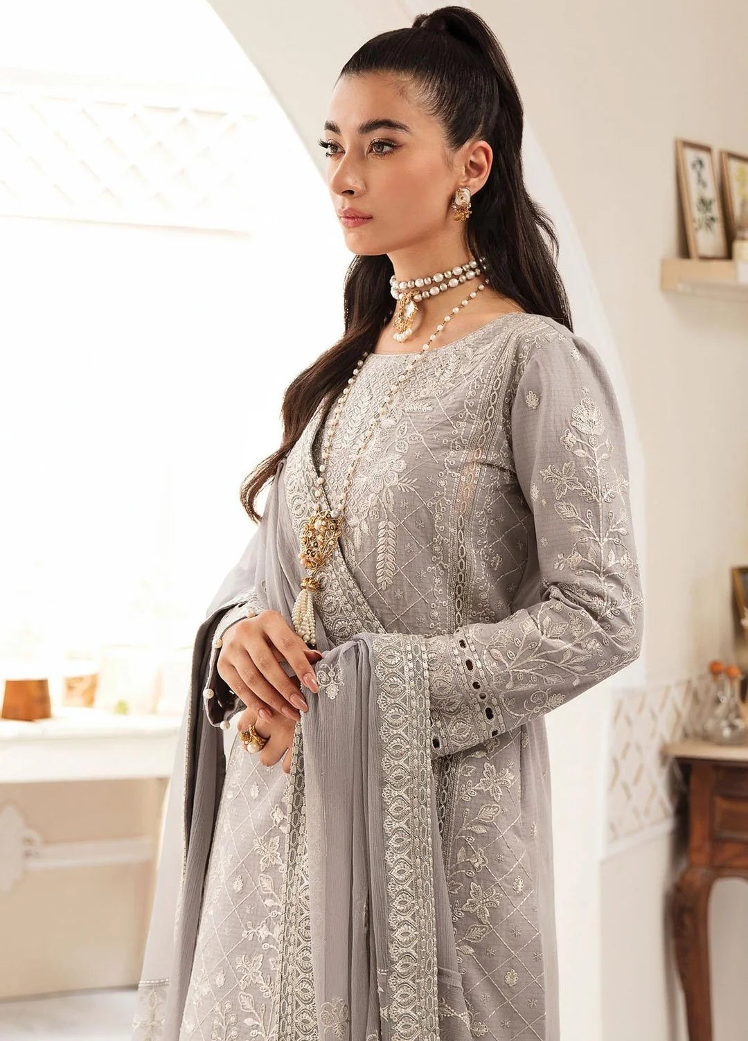 A woman wearing a light gray embroidered traditional outfit with a matching dupatta, standing in a bright indoor setting.