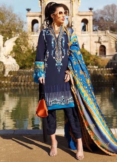 A woman models a traditional South Asian outfit consisting of a navy blue kurta with intricate gold and silver embroidery, matching navy pants, and a vibrant blue dupatta with gold and yellow patterns. She is standing outdoors near a body of water with a historic stone building in the background.