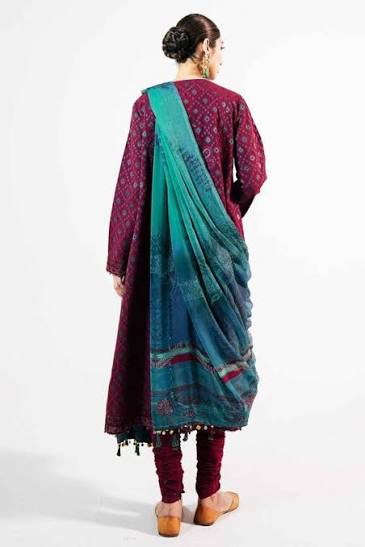 A woman shown from the back wearing a traditional three-piece salwar kameez suit. The kurta is deep magenta with intricate gold and blue patterns, paired with matching trousers and a teal-to-blue gradient dupatta draped over her shoulders.
