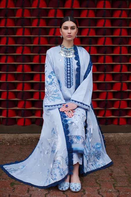 A woman wearing a long light blue embroidered traditional outfit with blue floral patterns and a matching dupatta, seated in front of a red and black patterned wall.