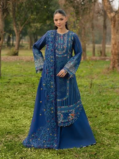 A woman stands outdoors on grass wearing a long blue embroidered traditional gown with ornate multicolor detailing and wide sleeves, photographed in a park-like setting with trees in the background.