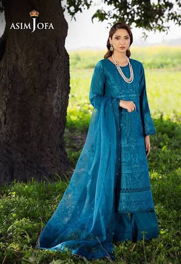 A woman wearing a teal blue embroidered traditional outfit with a matching sheer dupatta stands outdoors beside a large tree in a grassy field. The brand name ASIM JOFA is visible in the top left corner.