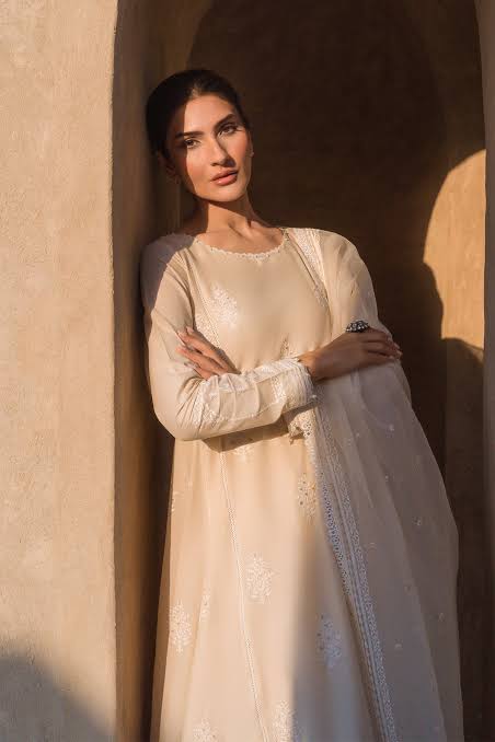 Woman in a cream traditional outfit standing against a textured wall.