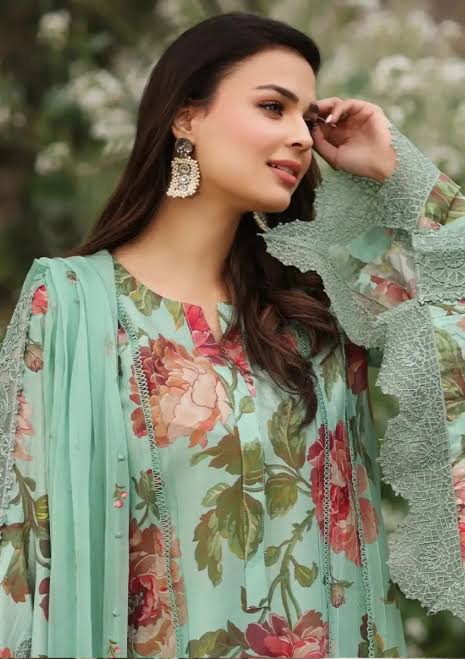 A woman wearing a mint green floral salwar kameez suit with lace sleeves and matching dupatta, styled with ornate gold earrings, set against a soft, blurred green background.