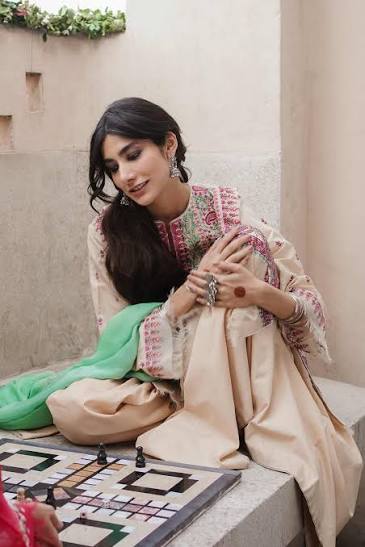 A woman in a beige embroidered traditional outfit with a green dupatta sits on a stone bench, looking down at a chessboard in a courtyard setting.