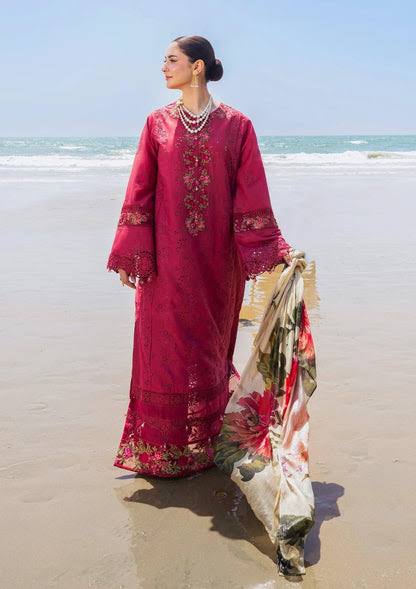 A woman stands on a sandy beach wearing a deep magenta embroidered traditional outfit with a matching floral dupatta draped over one arm. The background shows the ocean and clear sky.