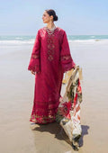 A woman stands on a sandy beach wearing a deep magenta embroidered traditional outfit with a matching floral dupatta draped over one arm. The background shows the ocean and clear sky.