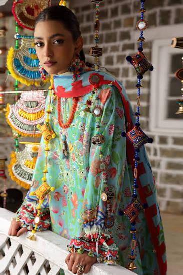 A woman wearing a highly decorated, colorful traditional long-sleeve top with intricate embroidery, beadwork, and metallic embellishments, posed in front of a rustic brick wall and hanging decorative ornaments.