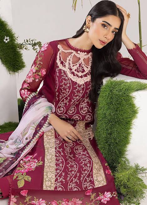 A woman models a deep maroon three-piece ethnic suit with intricate beige and pink floral embroidery, a matching dupatta, and a white embroidered inner kurta. She is posed in front of a white wall decorated with green foliage.