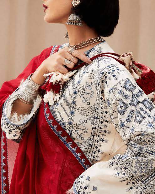A close-up of a model wearing traditional South Asian attire, featuring a cream-colored kurta with intricate blue and gray floral embroidery, paired with a rich red dupatta and matching red fabric.