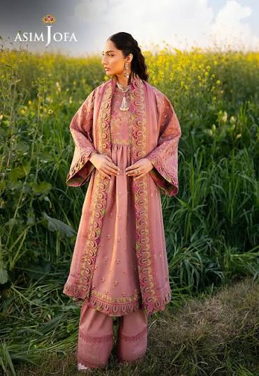 A woman stands outdoors in a field of yellow flowers wearing a dusty pink traditional outfit with ornate gold embroidery. The brand name 'ASIM JOFA' is visible in the top left corner.