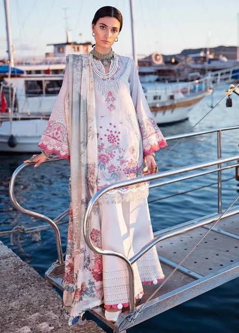 A woman models a traditional South Asian outfit with a light blue and white printed kurta, matching trousers, and a dupatta, standing on a metal gangway with a harbor and boats in the background.