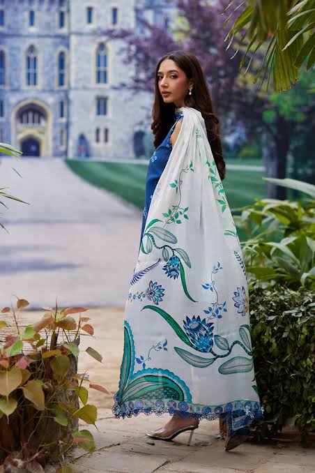 Woman in a blue top and white floral saree walking outdoors with a building and garden in the background