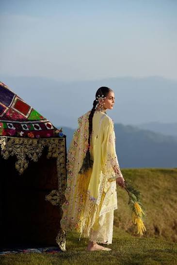 A woman in a pale yellow embroidered traditional outfit stands barefoot outdoors beside a decorated tent, holding a bundle of dried grass, with a hazy mountain landscape in the background.