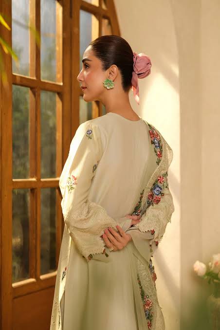 A woman in profile wearing a cream-colored traditional outfit with floral embroidery and a matching embroidered dupatta, standing in front of a warm, sunlit wooden window.
