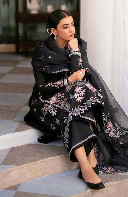 A woman sits on stone steps wearing a black embroidered traditional outfit with pink and white floral detailing, a matching sheer shawl, and black pointed shoes.