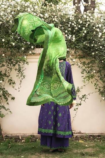 A person outdoors in front of a white wall covered with white flowers, wearing a purple traditional outfit with green border and pattern details, while holding a bright green patterned dupatta over their head.