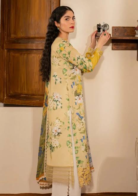 A woman wearing a long cream-colored floral printed kurta with yellow sleeves and white trousers, standing in an indoor setting and holding a vintage camera against a beige wall.