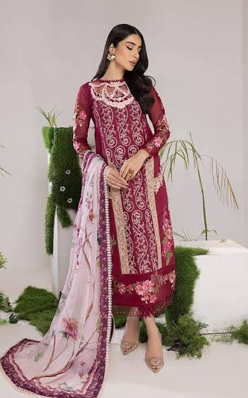 A woman models a traditional three-piece salwar kameez suit in deep maroon with intricate gold embroidery, paired with a light pink floral dupatta, set against a studio backdrop with decorative greenery.