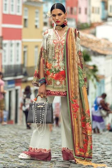 A model stands on a cobblestone street wearing a traditional South Asian outfit. The outfit features a cream-colored kurta with red and green floral patterns, wide-leg trousers in cream with red borders, and a matching dupatta with a gold and red floral print.