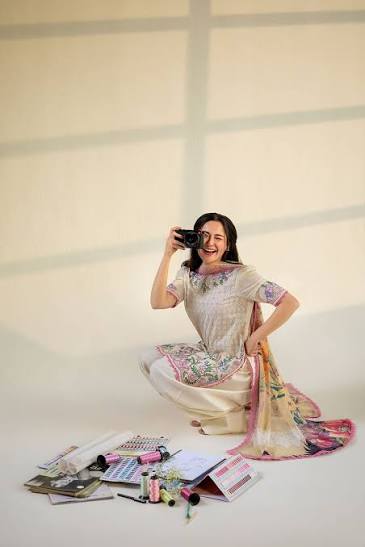 A woman in a light beige and pink floral printed traditional outfit kneels on a cream-colored floor, holding a black camera to her eye. Papers and small accessories are scattered on the floor in front of her.