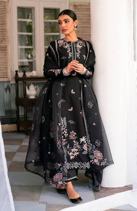 A woman models a long black embroidered dress with pink and white floral patterns, standing in a classic indoor setting with white columns.