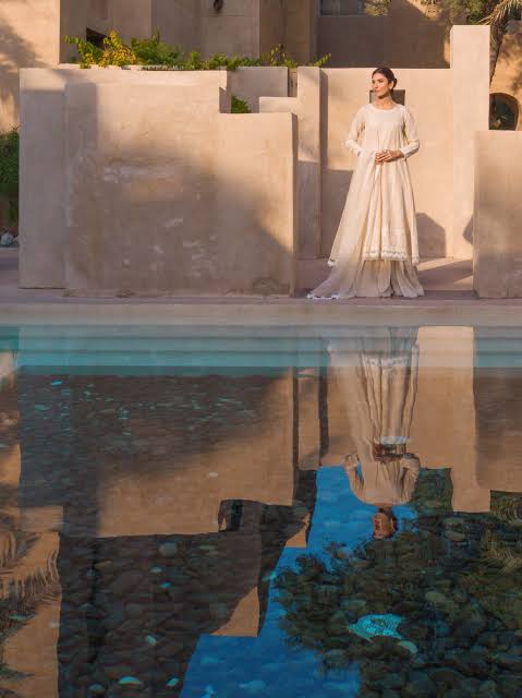 A woman wearing a long-sleeved cream-colored maxi dress stands beside a reflective pool, with her reflection clearly visible in the water. The background features warm beige stucco walls and architectural structures under warm sunlight.