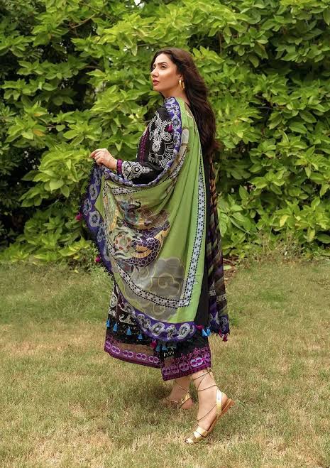 A woman wearing a green traditional outfit with purple and black patterned borders and a matching draped scarf, standing outdoors on grass in front of a leafy green hedge.