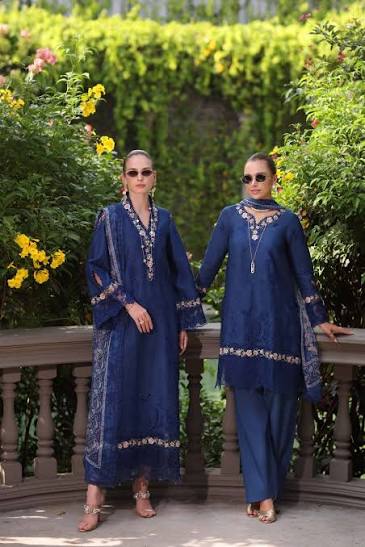 Two women modeling a navy blue ethnic three-piece suit with intricate gold embroidery, standing outdoors in front of a green hedge and stone railing.