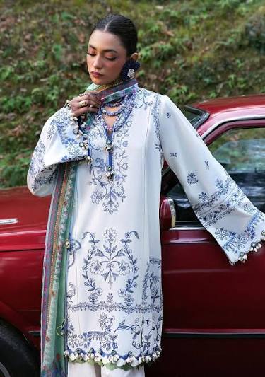 A woman wearing a white embroidered traditional outfit with blue floral patterns and a multicolored scarf, standing beside a red car outdoors.