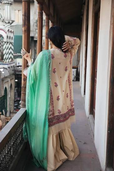 A woman viewed from behind wearing a traditional beige embroidered kurta with a green dupatta, standing on a wooden balcony.