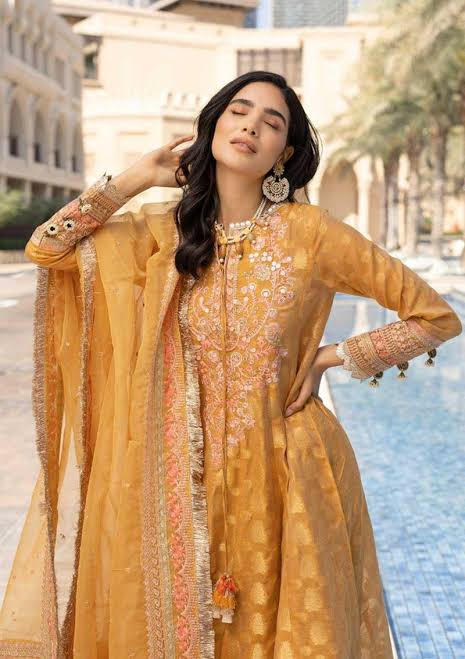 A woman modeling a traditional embroidered mustard yellow ethnic outfit with a matching sheer dupatta, standing outdoors in front of a blurred architectural background.