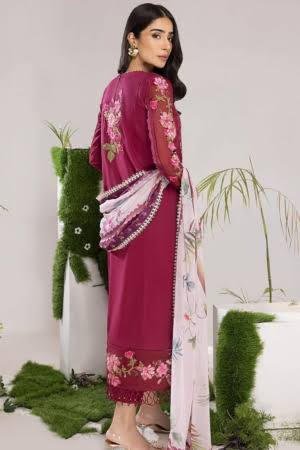 A woman models a three-piece traditional outfit featuring a magenta kurta with floral embroidery, a matching embroidered dupatta, and a light pink bottom skirt, set against a studio backdrop with decorative green plants.